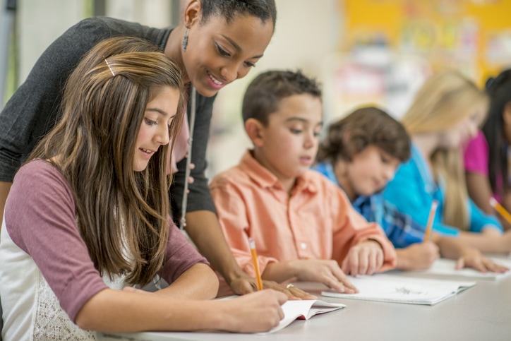 A multi-ethnic group of elementary children are sitting in class and are sitting at their desks are are taking a test. One student is getting some clarification on the test question.