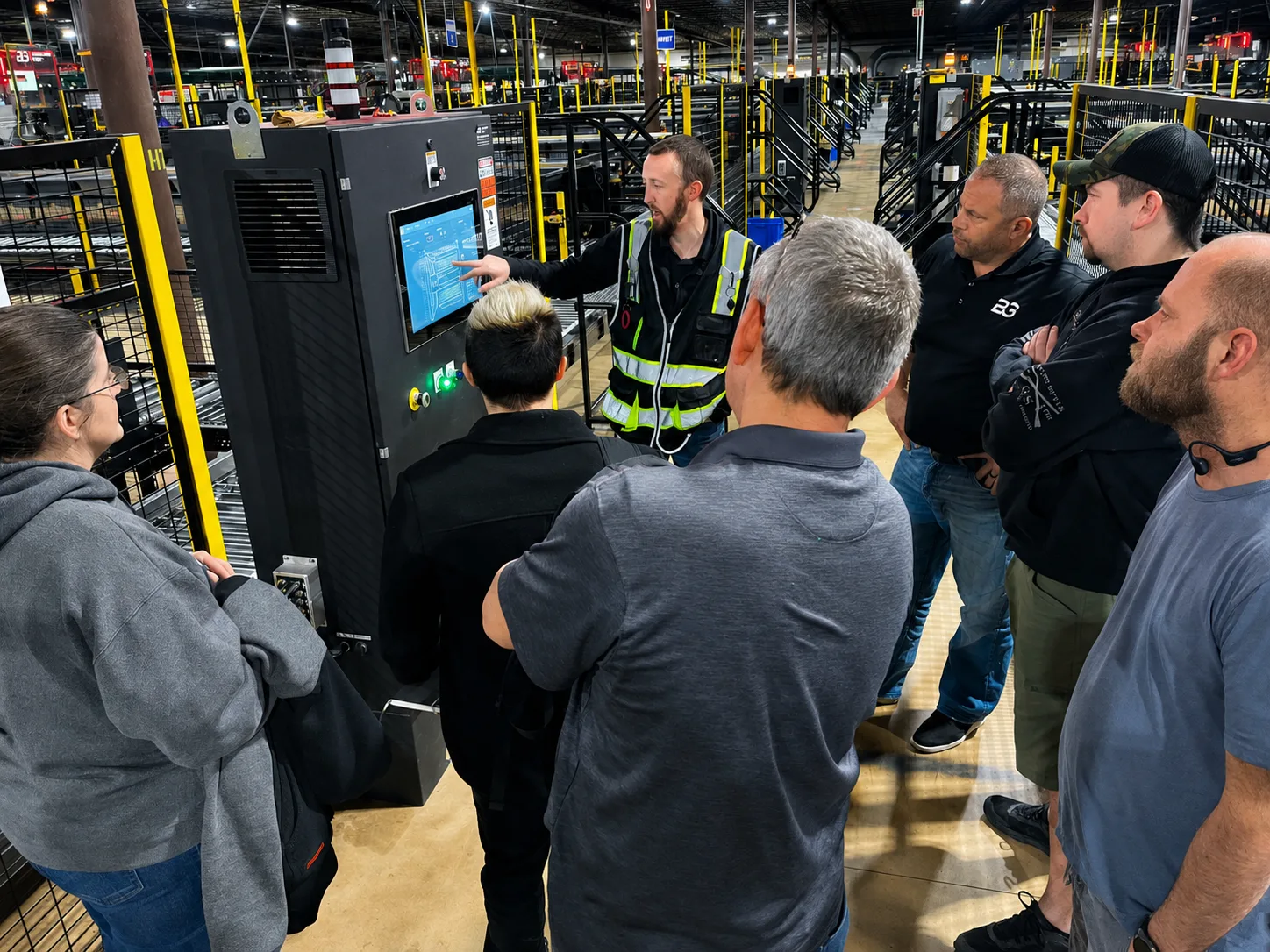 An instructor pointing at a touchscreen control panel while training a group of workers inside a warehouse facility.